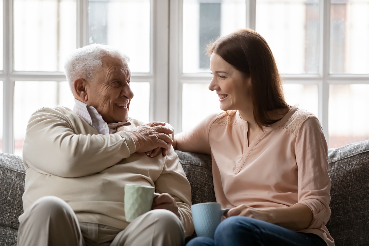 Elderly man and young woman smiling while sitting on a sofa, holding mugs